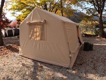 A large beige tent is set up in a leafy campsite surrounded by autumnal trees with colorful foliage. The ground is covered with small gravel stones and scattered fallen leaves. Two dark green canisters are placed near the entrance of the tent.