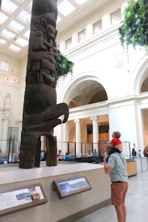 A large totem pole stands prominently in a brightly lit museum hall. The totem includes figures and animals carved from dark wood. A man with a child sitting on his shoulders is observing the totem. Surrounding the totem, there are informational displays and a few people sitting and walking in the background. The space is adorned with high ceilings, natural light from skylights, and hanging greenery.