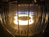 A close-up of a glowing heater element surrounded by a protective metal cage. The element emits a bright white and yellow light with hints of blue at the edges.