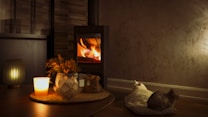 A cozy interior scene with a lit candle and decorative items on a wooden table in the foreground. In the background, a warm fire burns inside a black wood stove, casting a soft glow across the room. Two cats, one white and one gray, rest comfortably on a cushioned bed near the stove.