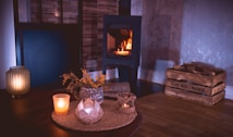 A cozy indoor setting featuring a lit fireplace surrounded by wood-paneled walls. In the foreground, a table holds a candle, a decorative jar with dried flowers, and other small items. To the right, a wooden crate filled with logs is placed on a polished wooden floor.