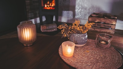 A cozy interior setting with a warm atmosphere. A lit candle in a glass holder stands on a woven mat alongside a ceramic vase containing yellow flowers. In the background, there is a wooden crate and a wood-burning stove with a glowing fire.