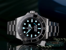 A luxury wristwatch with a luminous dial and markers, featuring a stainless steel bracelet and a rotating bezel. The watch face is adorned with the brand logo, offering a sleek and polished appearance against a dark background.