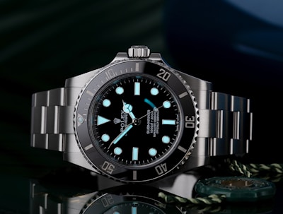A luxury wristwatch with a luminous dial and markers, featuring a stainless steel bracelet and a rotating bezel. The watch face is adorned with the brand logo, offering a sleek and polished appearance against a dark background.