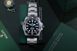 A luxury stainless steel watch with a rotating black bezel and luminous markers, placed on a dark green surface accompanied by a green warranty card and a manual. The watch face displays branding and details about its depth rating.