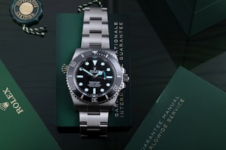A luxury stainless steel watch with a rotating black bezel and luminous markers, placed on a dark green surface accompanied by a green warranty card and a manual. The watch face displays branding and details about its depth rating.