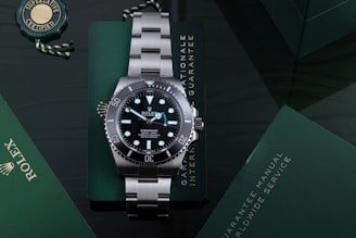 A luxury stainless steel watch with a rotating black bezel and luminous markers, placed on a dark green surface accompanied by a green warranty card and a manual. The watch face displays branding and details about its depth rating.