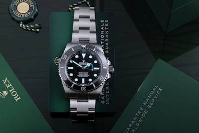 A luxury stainless steel watch with a rotating black bezel and luminous markers, placed on a dark green surface accompanied by a green warranty card and a manual. The watch face displays branding and details about its depth rating.
