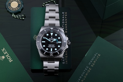 A luxury stainless steel watch with a rotating black bezel and luminous markers, placed on a dark green surface accompanied by a green warranty card and a manual. The watch face displays branding and details about its depth rating.