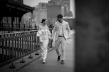 A couple dressed in elegant attire walks along a city sidewalk. The woman is wearing an off-shoulder white dress, while the man is in a light-colored suit. They are holding hands and smiling at each other, creating a sense of connection. The background shows an urban setting with buildings and a railing that runs parallel to their path.