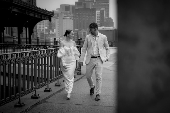 A couple dressed in elegant attire walks along a city sidewalk. The woman is wearing an off-shoulder white dress, while the man is in a light-colored suit. They are holding hands and smiling at each other, creating a sense of connection. The background shows an urban setting with buildings and a railing that runs parallel to their path.