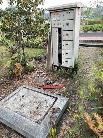 An electrical cabinet with multiple compartments stands in an outdoor setting. The cabinet is slightly open, revealing some wires inside. It's positioned near a small garden with various plants and a concrete slab lies in front of it, surrounded by dry leaves and debris. There are warning lights and gauges on the cabinet, suggesting it's in use.