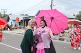 Two women in colorful attire stand by the roadside during a cultural event. The woman on the left is wearing a patterned headscarf and green top, smiling. The central figure is dressed in traditional attire, holding a pink umbrella and a decorative fan. In the background, there are people sitting and two more women in traditional costumes, one holding a red umbrella. The setting seems to be a street with houses and power lines visible behind them.
