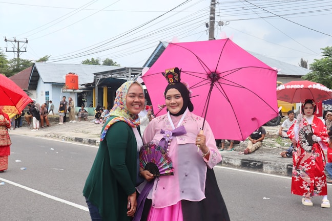 Two women in colorful attire stand by the roadside during a cultural event. The woman on the left is wearing a patterned headscarf and green top, smiling. The central figure is dressed in traditional attire, holding a pink umbrella and a decorative fan. In the background, there are people sitting and two more women in traditional costumes, one holding a red umbrella. The setting seems to be a street with houses and power lines visible behind them.