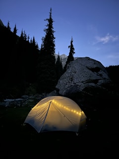A glowing tent illuminated from within is set against a mountainous backdrop with a darkening sky. Tall pine trees and a large boulder add to the serene wilderness setting.