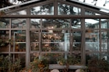 A greenhouse with a large glass facade showcasing various potted plants and gardening supplies. The structure, surrounded by lush greenery, has multiple panes and windows, allowing a clear view of the interior full of vibrant flora and organized shelves.