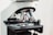A close-up of a microscope, focusing on the objective lenses. The lenses are metallic with engraved magnification details, surrounded by a black rotating disk. The microscope's stage is visible beneath the lenses with a holder mechanism for slides.