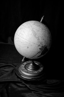 A monochrome globe showcasing various countries and continents, resting on a decorative stand with Roman numerals. The globe is set against a dark, draped fabric background.