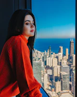 A stylish woman applying vibrant eyeshadow in front of a large urban window with skyline views.