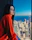 Photo of a cheerful woman in business casual attire standing near a window with city views.