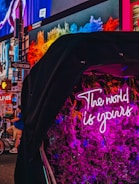A vibrant urban scene featuring a neon sign reading 'The world is yours' surrounded by pink flowers. In the background, colorful digital billboards display various advertisements in a bustling city environment with visible street signs and a person on a bicycle.