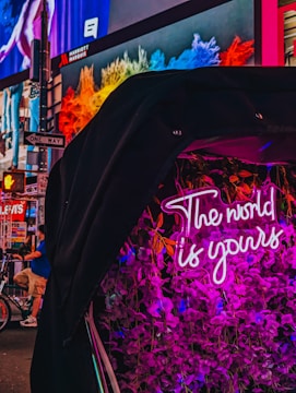 A vibrant urban scene featuring a neon sign reading 'The world is yours' surrounded by pink flowers. In the background, colorful digital billboards display various advertisements in a bustling city environment with visible street signs and a person on a bicycle.