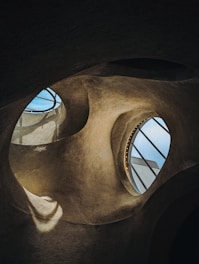Curved architectural shapes with large, circular openings reveal glimpses of the sky. The textured surfaces and organic forms create a cave-like or futuristic interior with light streaming through the openings.