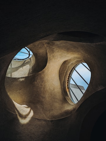 Curved architectural shapes with large, circular openings reveal glimpses of the sky. The textured surfaces and organic forms create a cave-like or futuristic interior with light streaming through the openings.