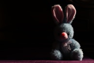 A plush blue bunny with pink inner ears and a pink nose is positioned against a dark background. The bunny is wearing a sheer ribbon tied around its neck and is sitting in a spotlight that highlights its fluffy texture, creating a strong contrast with the surrounding shadows.
