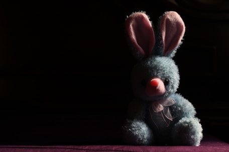 A plush blue bunny with pink inner ears and a pink nose is positioned against a dark background. The bunny is wearing a sheer ribbon tied around its neck and is sitting in a spotlight that highlights its fluffy texture, creating a strong contrast with the surrounding shadows.