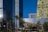 Tall, modern skyscrapers tower above the landscape with a mix of geometric glass and metallic designs. Palm trees line the foreground, adding a touch of greenery to the urban setting. Large, prominent signs for luxury brands Gucci and Dolce & Gabbana contrast with the sleek buildings.