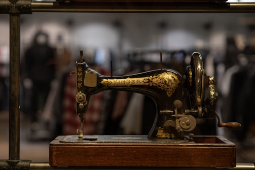 An antique sewing machine with intricate gold detailing on its black body, positioned prominently in the foreground. The sewing machine sits on a wooden base, and a blurred background reveals racks of clothing, suggesting a vintage or nostalgic setting.