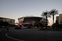 A large modern arena with the name T-Mobile Arena prominently displayed on its roof. The structure is surrounded by palm trees and various other buildings in an urban setting. Vehicles are visible on the road in front of the arena, with a few pedestrians nearby. The sky is dusky, suggesting the time is either early morning or late evening.