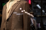 A mannequin is dressed in a brown textured suit jacket over a light patterned shirt. The image is taken through a glass, creating reflections and adding depth. The background is slightly blurred, with hints of industrial and modern elements.