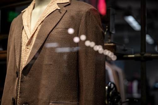 A mannequin is dressed in a brown textured suit jacket over a light patterned shirt. The image is taken through a glass, creating reflections and adding depth. The background is slightly blurred, with hints of industrial and modern elements.
