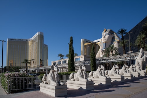 A large hotel complex with a distinct Egyptian theme features a prominent statue of a pharaoh's head, flanked by palm trees and sphinx sculptures. The buildings have a modern architectural style, and the golden Mandalay Bay hotel is prominently visible in the background.