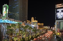 A bustling city street at night with bright neon lights illuminating numerous buildings, including hotels and casinos. The scene is lively with heavy traffic on the road and palm trees lining the street. Large digital billboards display advertisements.