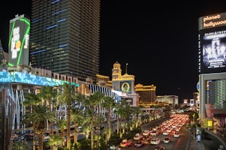 A bustling city street at night with bright neon lights illuminating numerous buildings, including hotels and casinos. The scene is lively with heavy traffic on the road and palm trees lining the street. Large digital billboards display advertisements.