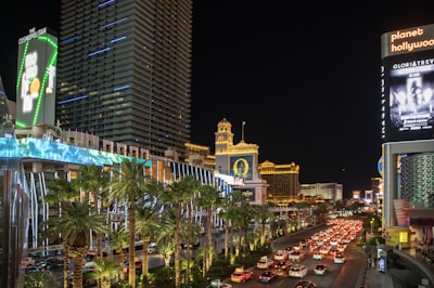 A bustling city street at night with bright neon lights illuminating numerous buildings, including hotels and casinos. The scene is lively with heavy traffic on the road and palm trees lining the street. Large digital billboards display advertisements.