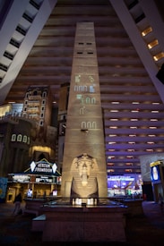 A massive indoor atrium features an obelisk adorned with Egyptian hieroglyphs, leading to a large statue of a pharaoh. The surrounding area includes hotel interiors with multiple floors visible, and illuminated signs for an atrium showroom and food court.