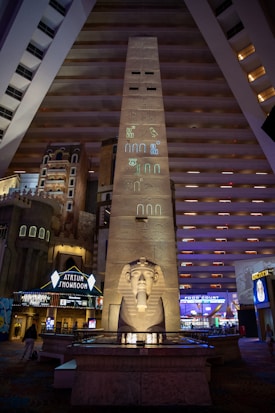 A massive indoor atrium features an obelisk adorned with Egyptian hieroglyphs, leading to a large statue of a pharaoh. The surrounding area includes hotel interiors with multiple floors visible, and illuminated signs for an atrium showroom and food court.