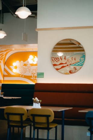 A modern cafe interior featuring colorful wall art and round light fixtures. The seating includes booths with orange and dark green cushions and wooden tables with chairs. A circular mirror reflects part of the art, which depicts stylized landscapes.