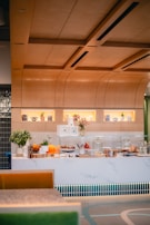 A modern cafe counter featuring a variety of culinary displays with pastries, cakes, and fresh fruits. The counter is adorned with plants and decorative items. Warm lighting and wooden accents create a cozy atmosphere.