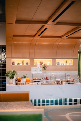 A modern cafe counter featuring a variety of culinary displays with pastries, cakes, and fresh fruits. The counter is adorned with plants and decorative items. Warm lighting and wooden accents create a cozy atmosphere.