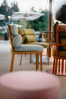 An outdoor seating area featuring modern chairs with rounded, cushioned backs and slender frames. The chairs are green and orange, and there's a soft-focus effect highlighting the atmosphere. In the background, large white umbrellas provide shade, and lush greenery is visible.