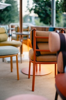 A collection of modern chairs and small tables placed on a patio, surrounded by greenery. The chairs are upholstered in various colors, with bright orange frames and subtle earth-toned cushions. The atmosphere is relaxed and inviting.