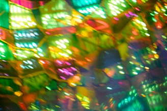 A vibrant, multicolored abstract pattern featuring overlapping geometric shapes with intense hues of green, yellow, orange, pink, and blue. The colors appear luminous and blend into each other, creating a kaleidoscopic effect.