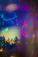 An abstract composition featuring a blend of colorful lights and shadows, with outlines of trees visible against a backdrop of vibrant hues. The image includes a mixture of deep blues, bright pinks, and soft yellows creating a dreamlike atmosphere.