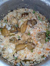 A large pot filled with a dish consisting of cooked rice mixed with pieces of cooked meat. The rice is seasoned and colored, likely with spices and herbs, showing hints of orange and green. Visible pieces of meat are interspersed throughout the rice, and some garnishing with herbs can be spotted.