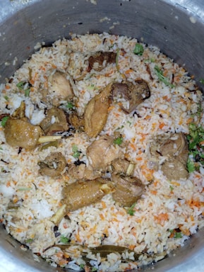 A large pot filled with a dish consisting of cooked rice mixed with pieces of cooked meat. The rice is seasoned and colored, likely with spices and herbs, showing hints of orange and green. Visible pieces of meat are interspersed throughout the rice, and some garnishing with herbs can be spotted.
